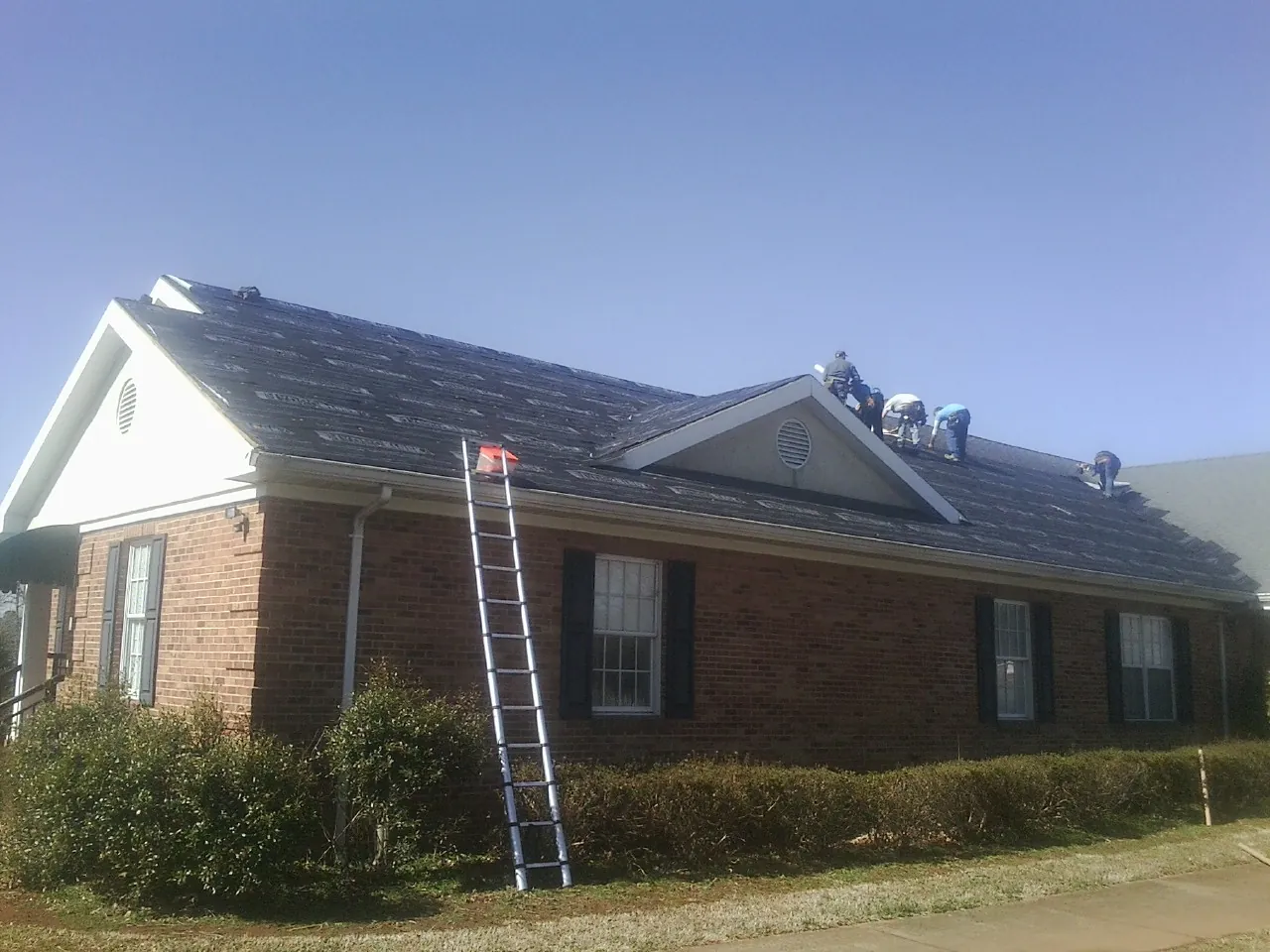 Crew tearing shingles off a brick ranch roof with a ladder set — Metal Roof Repair work by our crew in Milton Mills
