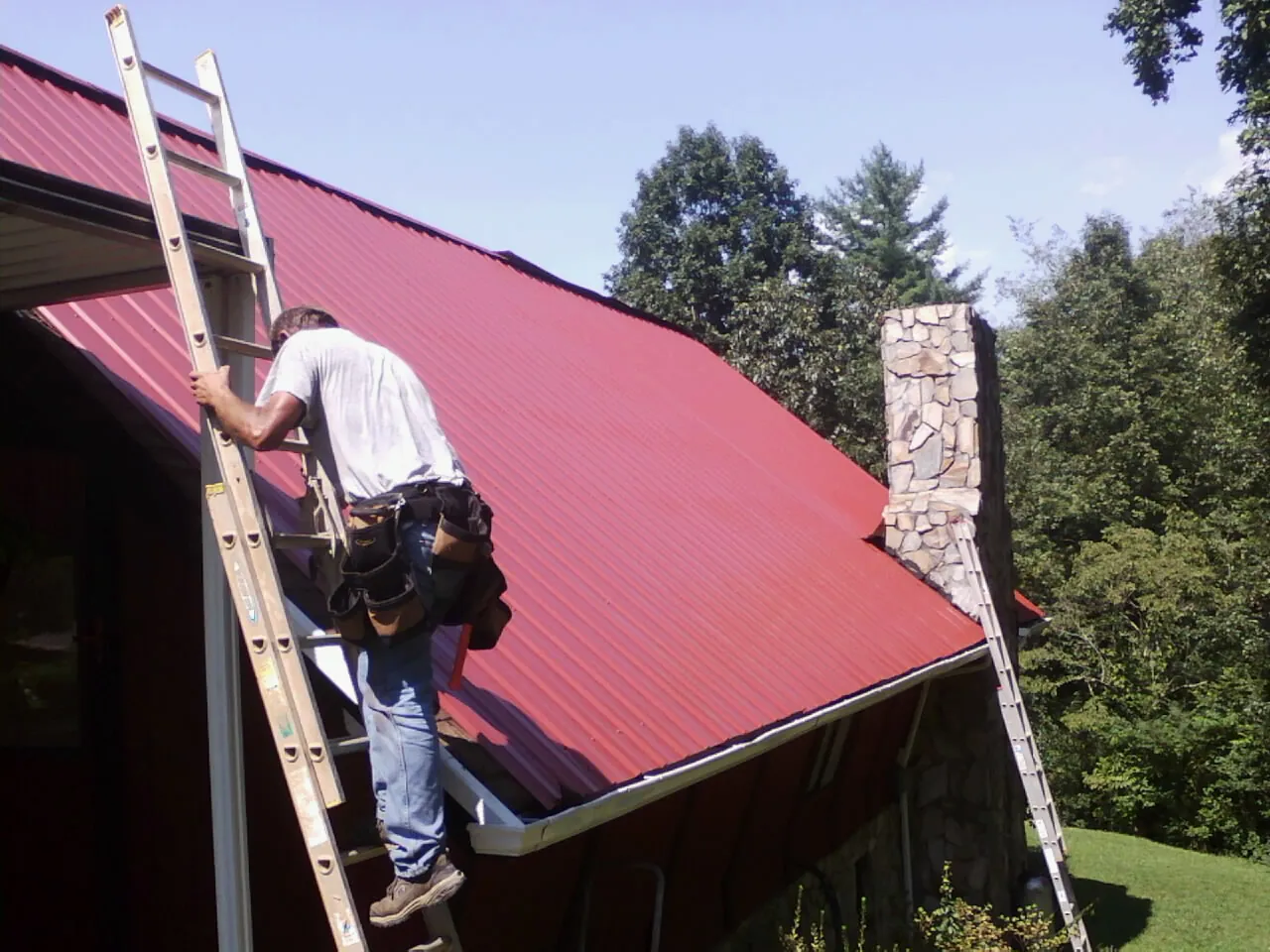 Roofer on a ladder beside a red metal roof and stone chimney — Soffit & Fascia Repair by Roofing Brothers in Milton Mills
